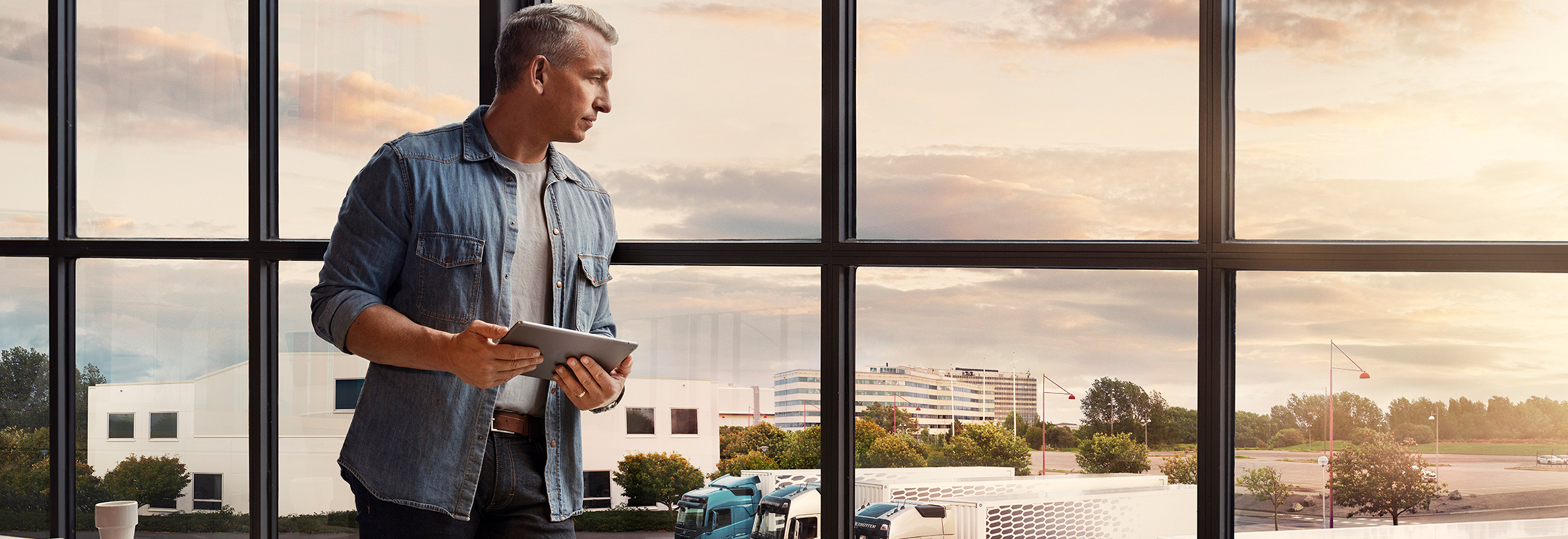 Ένας άντρας που κρατά ένα tablet στέκεται δίπλα σε ένα παράθυρο και ατενίζει το στόλο των φορτηγών
